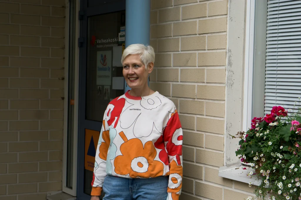 Toimitaax Oy:n sisäänkäynti. Toimintaterapeutti Jaana Juhola-Lahtinen seisoo oven vieressä, ja ovessa on Toimitaax Oy:n kyltti.